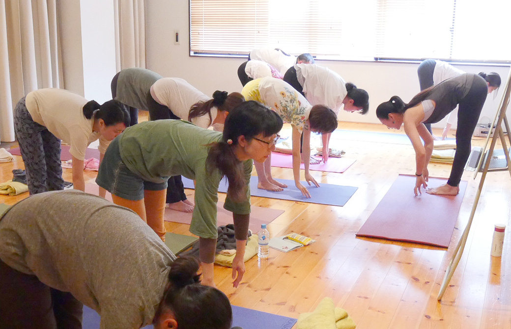 ヨガ講師(磯部佳世子)に習って生徒20名ほどが一緒に立位前屈をしている様子