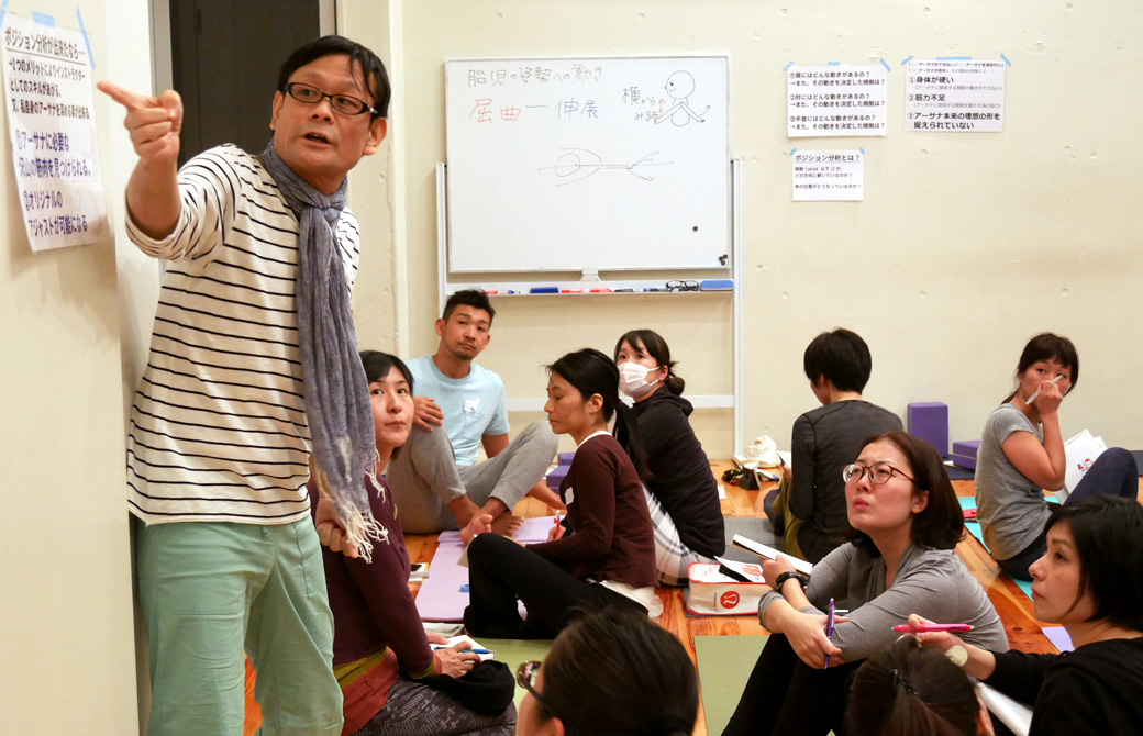ヨガ解剖学講座基礎編の講座風景。講師(内田かつのり)が壁に貼った資料を指差しながら説明し、それを真剣にきいている生徒の様子